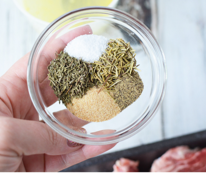 Small glass bowl being held above steaks filled with spices for steak marinade