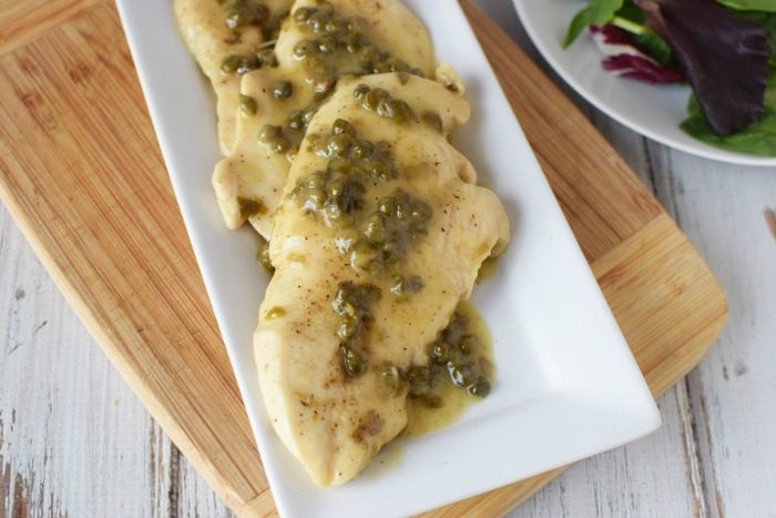 Chicken piccata laying on a white platter that is sitting on a wooden cutting board and topped with sauce and capers