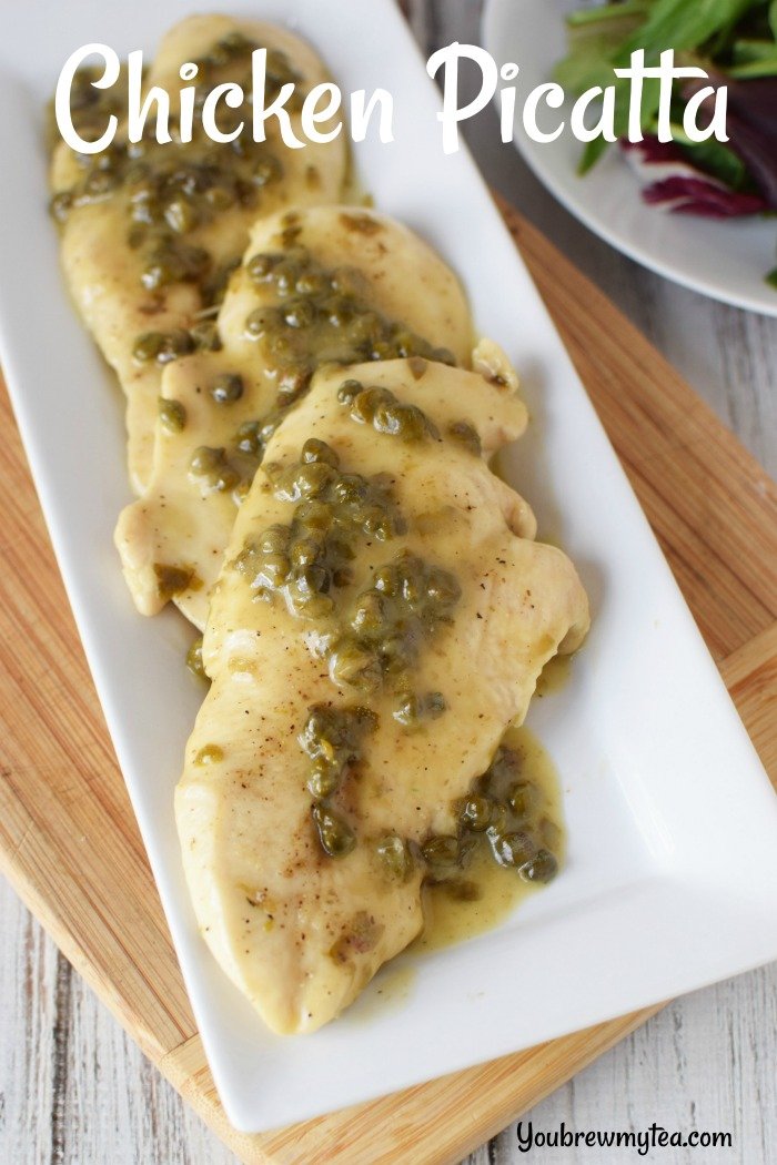 Chicken piccata laying on a white platter that is sitting on a wooden cutting board and topped with sauce and capers