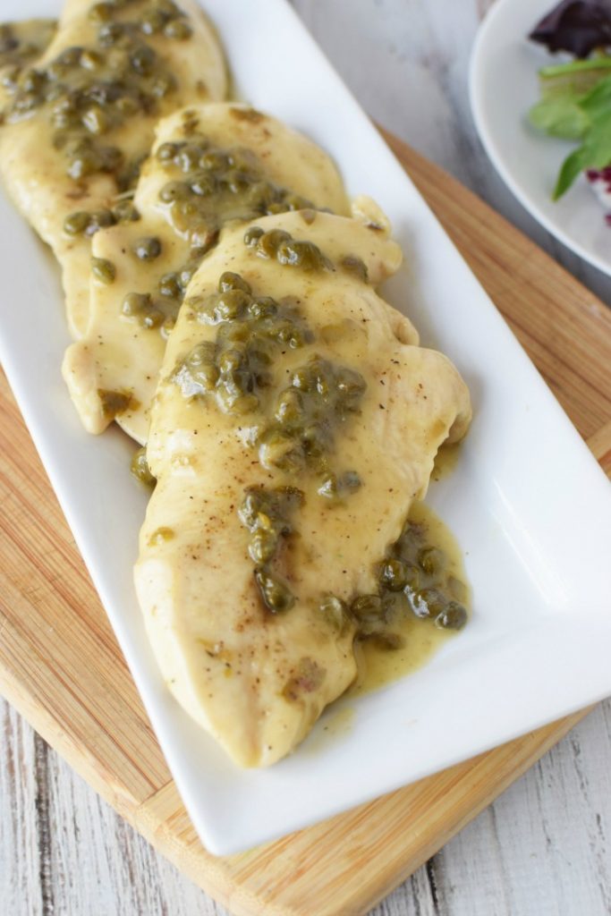 Chicken piccata laying on a white platter that is sitting on a wooden cutting board and topped with sauce and capers