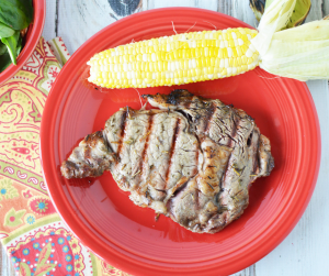 Ribeye steak grilled and laying on a red plate alongside an ear of corn that is also sitting on a colorful napkin
