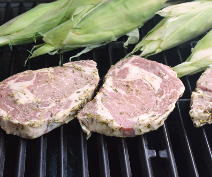 2 ribeye steaks on a grill alongside whole ears of corn