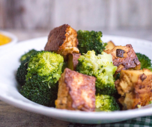 A large white plate filled with broccoli and crispy tofu sitting on a table with a small bowl of mango sauce in the background