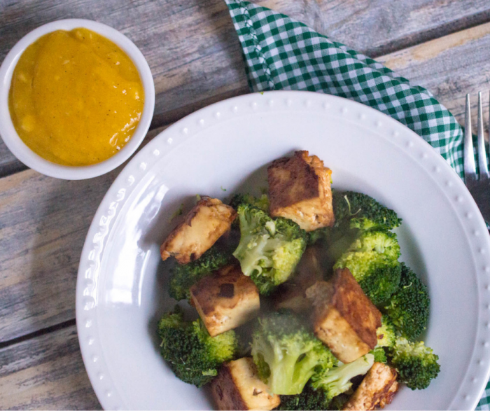 Overhead picture of a white bowl of crispy tofu and broccoli laying on a wooden surface with a small ramekin of mango sauce to the side and a blue checkered napkin and fork tucked beside the bowl