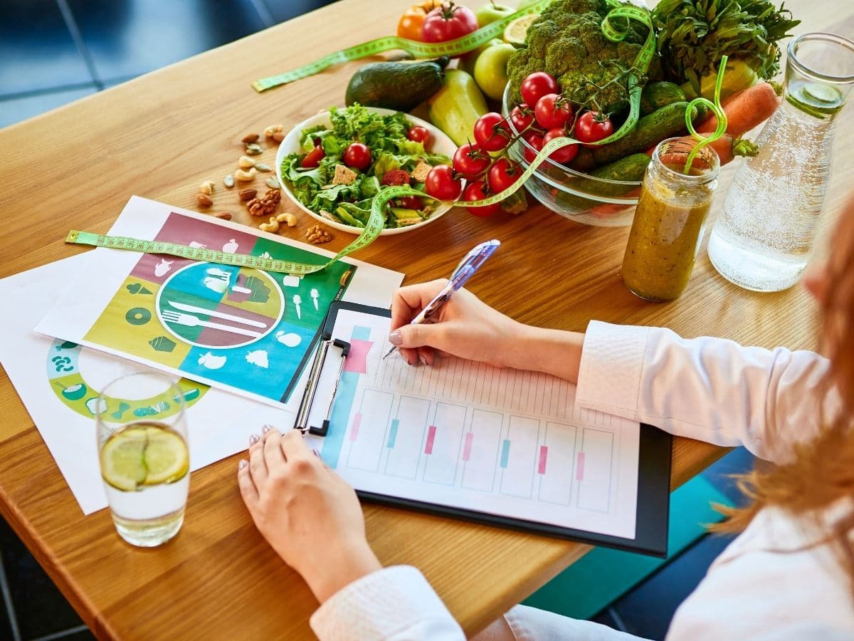 woman at desk with printed meal plan and food chart next to bowls of vegetables