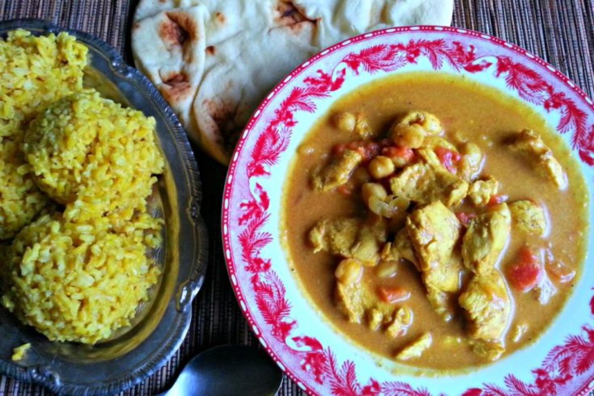 curry in red and white bowl next to bowl of rice and naan