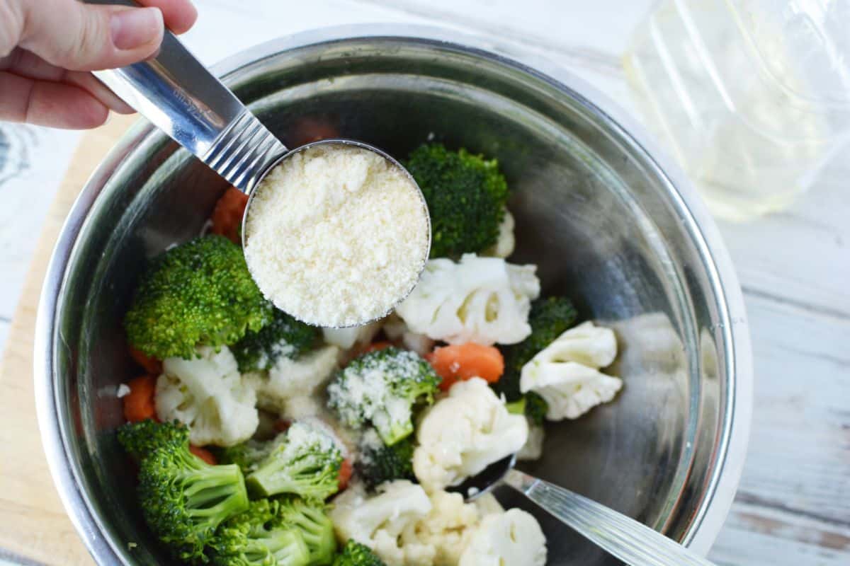 measuring cup of parmesan held above stainless steel bowl of vegetables